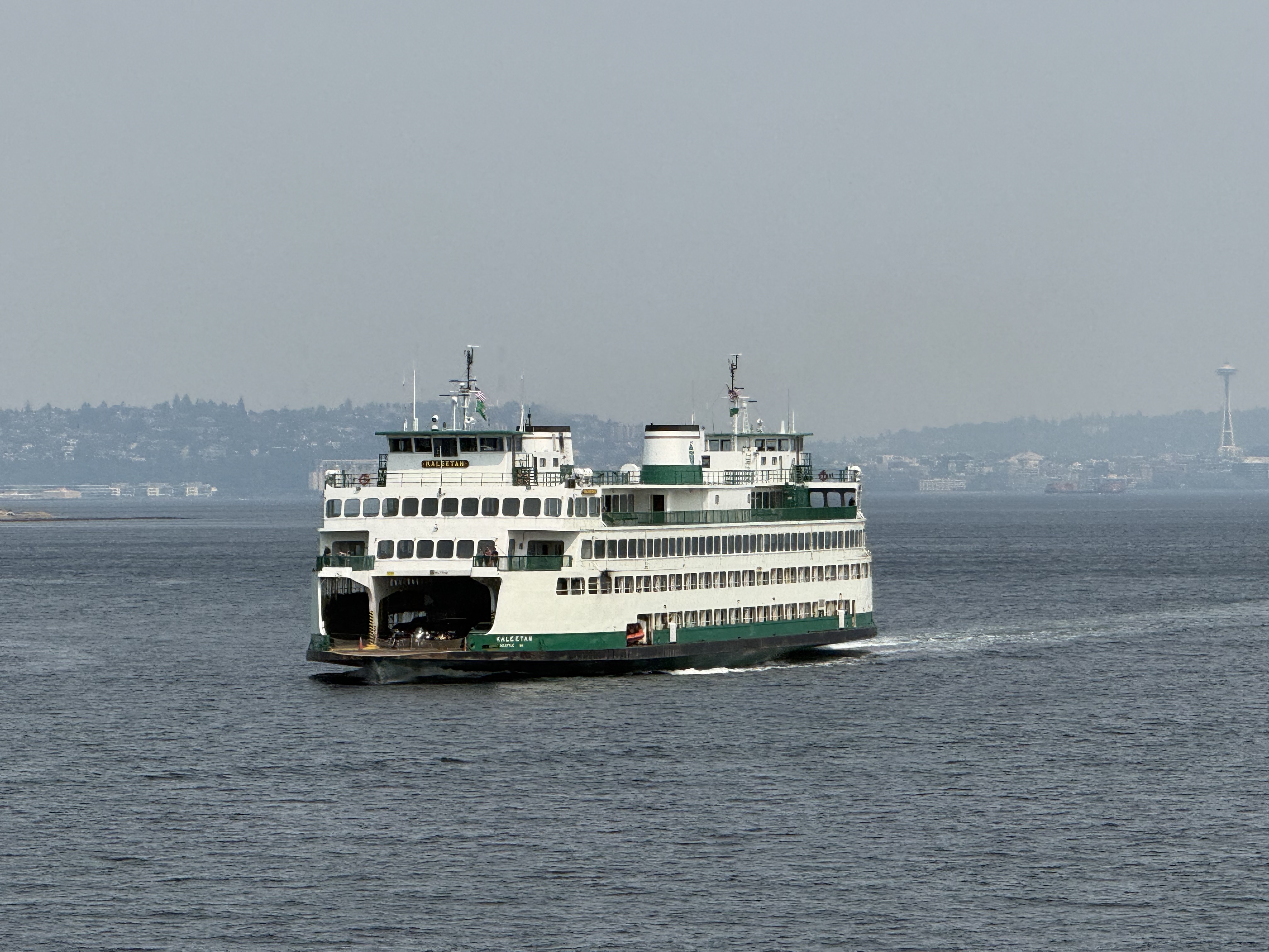 A large ferry boat navigating through calm waters with the Seattle skyline in the background, including the Space Needle.